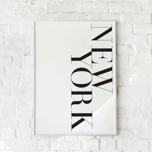 K&ouml;nnte beinhalten: Ein wei&szlig;er gerahmter Druck mit dem Wort "NEW YORK" in schwarzen Buchstaben, der an einer wei&szlig;en Backsteinwand h&auml;ngt.