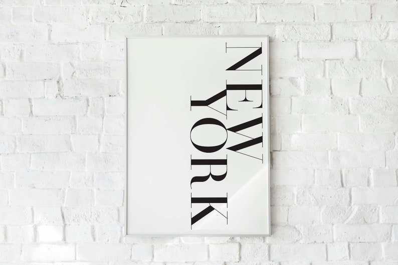 K&ouml;nnte beinhalten: Ein wei&szlig;er gerahmter Druck mit dem Wort "NEW YORK" in schwarzen Buchstaben, der an einer wei&szlig;en Backsteinwand h&auml;ngt.