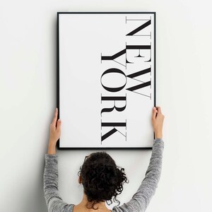 K&ouml;nnte beinhalten: Eine Person h&auml;ngt einen schwarzen gerahmten Druck an einer wei&szlig;en Wand. Der Druck zeigt das Wort "NEW YORK" in einer fetten, stilisierten Schrift.