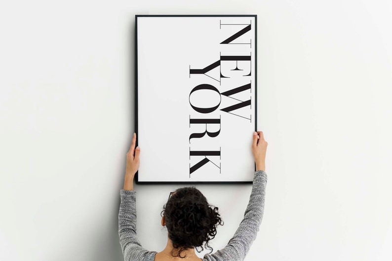 K&ouml;nnte beinhalten: Eine Person h&auml;ngt einen schwarzen gerahmten Druck an einer wei&szlig;en Wand. Der Druck zeigt das Wort "NEW YORK" in einer fetten, stilisierten Schrift.