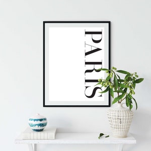 K&ouml;nnte beinhalten: Ein schwarzer gerahmter Druck mit dem Wort "PARIS" in wei&szlig;en Buchstaben auf wei&szlig;em Hintergrund. Der Druck steht auf einem wei&szlig;en Regal mit einer blau-wei&szlig;en Keramikvase und einer gr&uuml;nen Pflanze.