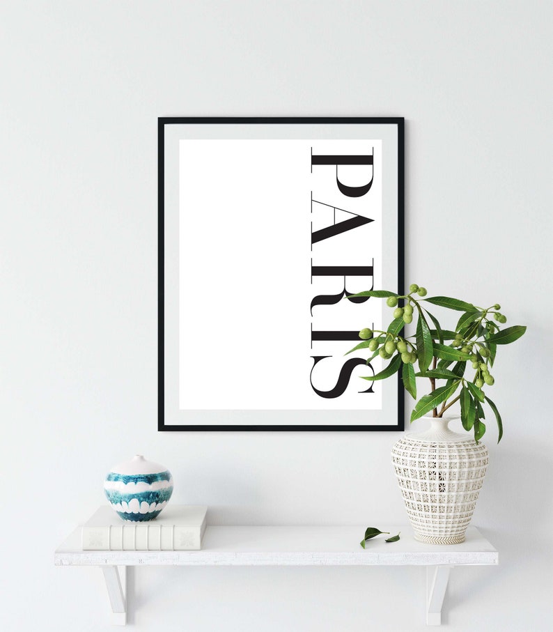 K&ouml;nnte beinhalten: Ein schwarzer gerahmter Druck mit dem Wort "PARIS" in wei&szlig;en Buchstaben auf wei&szlig;em Hintergrund. Der Druck steht auf einem wei&szlig;en Regal mit einer blau-wei&szlig;en Keramikvase und einer gr&uuml;nen Pflanze.