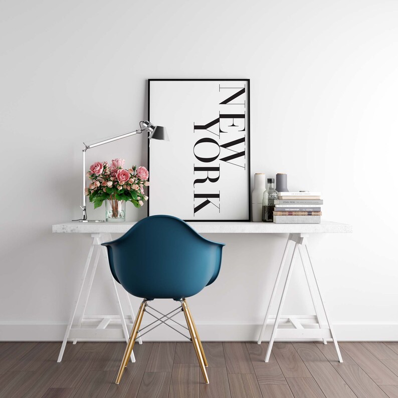 K&ouml;nnte beinhalten: Ein wei&szlig;er Schreibtisch mit einem wei&szlig;en Bockgestell, ein blauer Stuhl mit goldenen Beinen, ein schwarzer gerahmter Druck mit dem Wort "NEW YORK" in Wei&szlig;, eine Vase mit rosa Blumen und eine Lampe auf dem Schreibtisch.