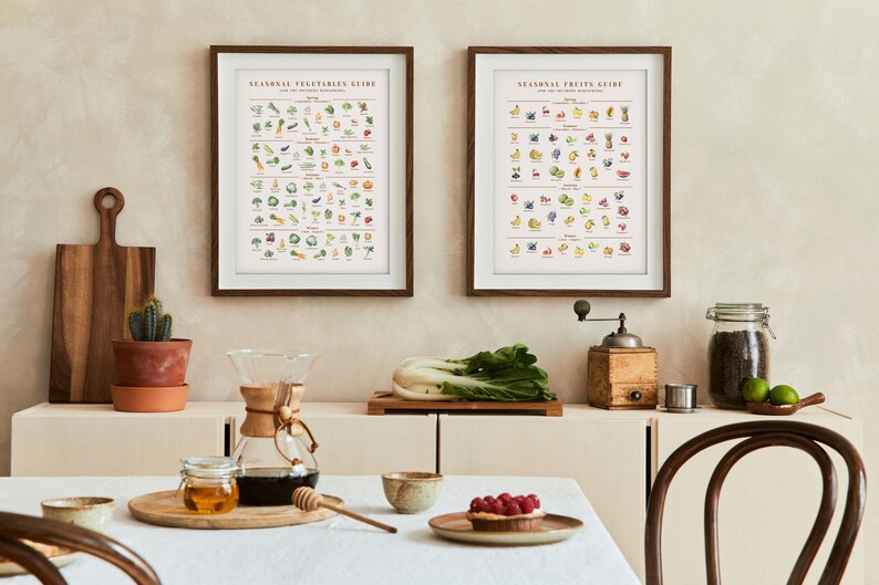 Puede incluir: Dos impresiones enmarcadas, "Gu&iacute;a de verduras de temporada" y "Gu&iacute;a de frutas de temporada", cuelgan sobre un armario blanco. Se ven una tabla de cortar de madera, un cactus en maceta de terracota y una cafetera Chemex. Una mesa est&aacute; puesta con una tarta y miel.