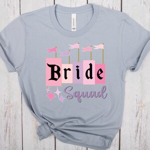 May include: A light blue t-shirt with the text "Bride Squad" in black lettering. The word "Bride" is in pink and purple blocks with gold trim. There are pink and purple flags above the word "Bride".