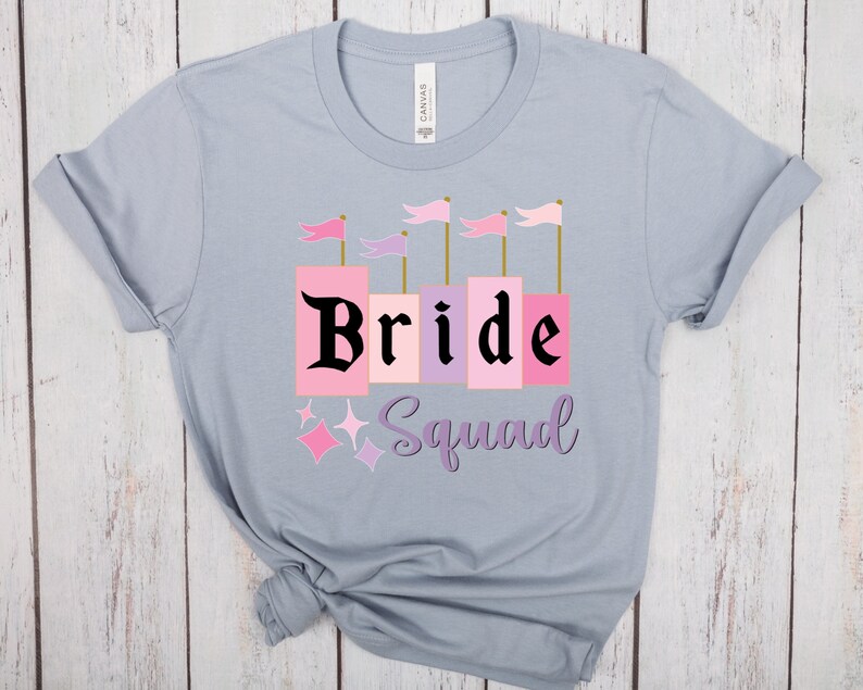 May include: A light blue t-shirt with the text "Bride Squad" in black lettering. The word "Bride" is in pink and purple blocks with gold trim. There are pink and purple flags above the word "Bride".