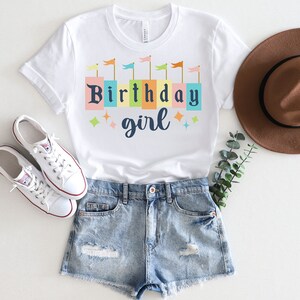 Peut inclure: T-shirt blanc avec un motif color&eacute; de fille d'anniversaire. Le motif repr&eacute;sente une rang&eacute;e de blocs color&eacute;s avec les mots "Birthday girl" et des drapeaux au-dessus des blocs.