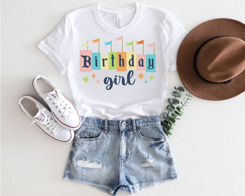 Peut inclure: T-shirt blanc avec un motif color&eacute; de fille d'anniversaire. Le motif repr&eacute;sente une rang&eacute;e de blocs color&eacute;s avec les mots "Birthday girl" et des drapeaux au-dessus des blocs.