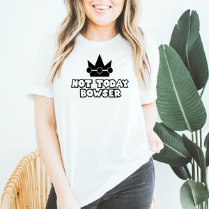 Puede incluir: Camiseta blanca con una corona negra y el texto "NOT TODAY BOWSER".