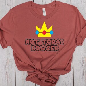 Puede incluir: Una camiseta marr&oacute;n con una corona amarilla, c&iacute;rculos azules y rojos y el texto "NOT TODAY BOWSER" en negro.
