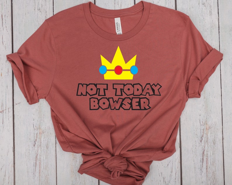 Puede incluir: Una camiseta marr&oacute;n con una corona amarilla, c&iacute;rculos azules y rojos y el texto "NOT TODAY BOWSER" en negro.