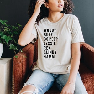 K&ouml;nnte beinhalten: Ein beiges T-Shirt mit schwarzem Text, der "Woody Buzz Bo Peep Jessie Rex Slinky Hamm" lautet.