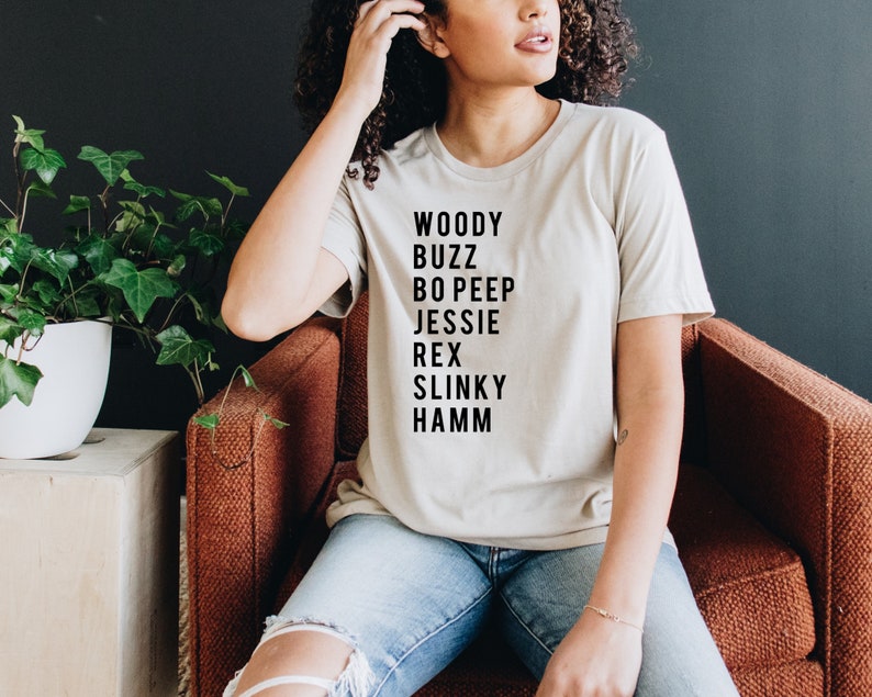 K&ouml;nnte beinhalten: Ein beiges T-Shirt mit schwarzem Text, der "Woody Buzz Bo Peep Jessie Rex Slinky Hamm" lautet.