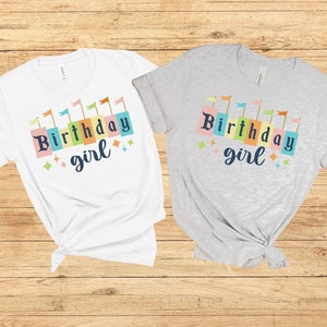 Peut inclure: Deux t-shirts avec le texte "Birthday girl" en lettres majuscules color&eacute;es avec des drapeaux au-dessus du texte. Un t-shirt est blanc et l'autre est gris.