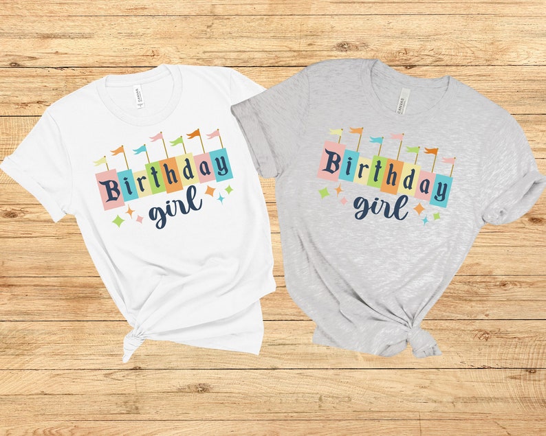 Peut inclure: Deux t-shirts avec le texte "Birthday girl" en lettres majuscules color&eacute;es avec des drapeaux au-dessus du texte. Un t-shirt est blanc et l'autre est gris.