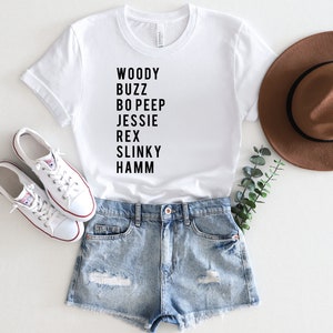 K&ouml;nnte beinhalten: Wei&szlig;es T-Shirt mit schwarzem Text, der "Woody Buzz Bo Peep Jessie Rex Slinky Hamm" lautet. Das Shirt wird mit Jeans-Shorts, wei&szlig;en Turnschuhen und einem braunen Fedora kombiniert.