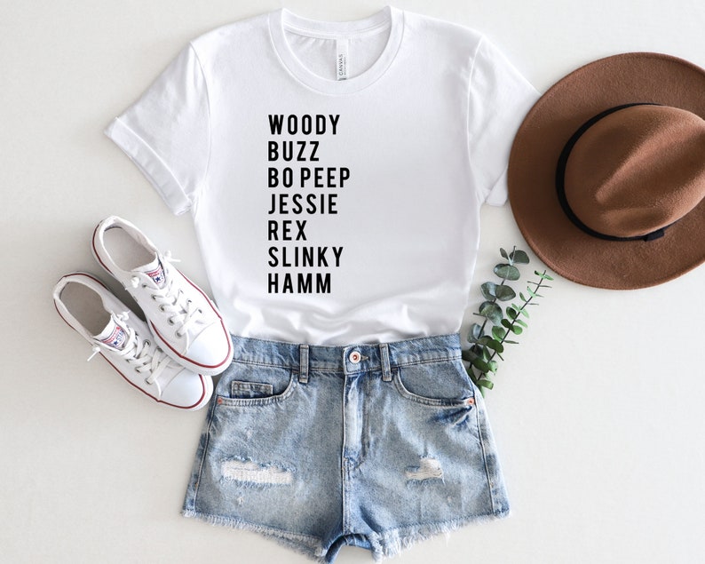 K&ouml;nnte beinhalten: Wei&szlig;es T-Shirt mit schwarzem Text, der "Woody Buzz Bo Peep Jessie Rex Slinky Hamm" lautet. Das Shirt wird mit Jeans-Shorts, wei&szlig;en Turnschuhen und einem braunen Fedora kombiniert.
