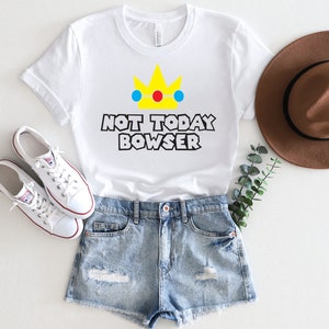 Puede incluir: Camiseta blanca con una corona amarilla y el texto "Not Today Bowser" en negro. La camiseta se combina con pantalones cortos de mezclilla y zapatillas blancas.