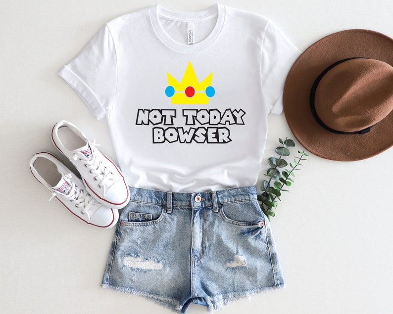 Puede incluir: Camiseta blanca con una corona amarilla y el texto "Not Today Bowser" en negro. La camiseta se combina con pantalones cortos de mezclilla y zapatillas blancas.