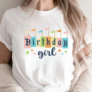 Peut inclure: T-shirt blanc avec un motif color&eacute; de fille d'anniversaire. Le motif comprend une rang&eacute;e de drapeaux color&eacute;s et les mots "Birthday girl" dans une police de caract&egrave;res en blocs.