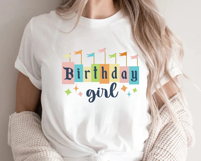 Peut inclure: T-shirt blanc avec un motif color&eacute; de fille d'anniversaire. Le motif comprend une rang&eacute;e de drapeaux color&eacute;s et les mots "Birthday girl" dans une police de caract&egrave;res en blocs.