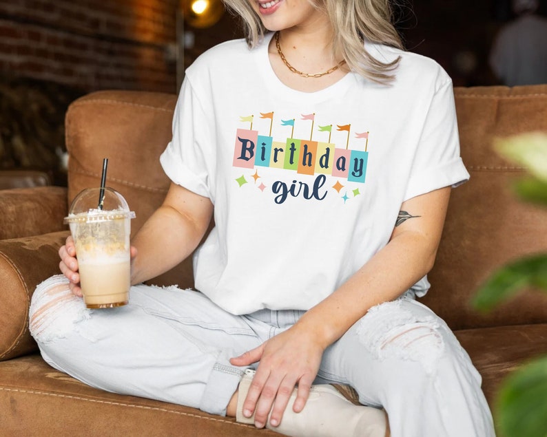 Peut inclure: Un t-shirt blanc avec un motif color&eacute; qui dit "Birthday girl" avec des drapeaux et des &eacute;toiles autour du texte.