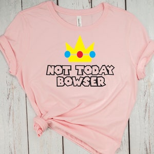 Puede incluir: Camiseta rosa con una corona amarilla, detalles azules y rojos, y el texto "Not Today Bowser" en negro.