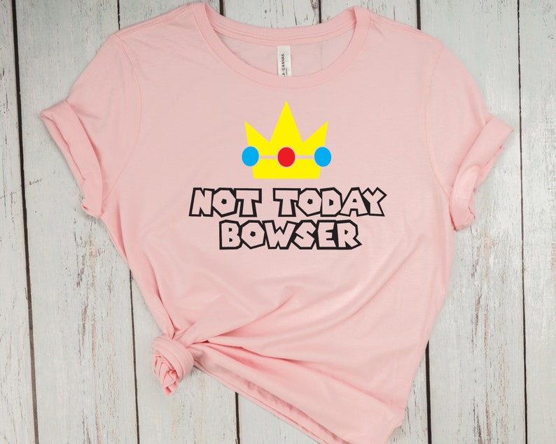 Puede incluir: Camiseta rosa con una corona amarilla, detalles azules y rojos, y el texto "Not Today Bowser" en negro.