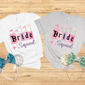 May include: Two white t-shirts with the text "Bride Squad" in pink and purple letters. The shirts are on a wooden background with a pair of sparkly mouse ears on each side.