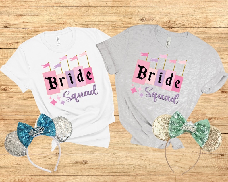 May include: Two white t-shirts with the text "Bride Squad" in pink and purple letters. The shirts are on a wooden background with a pair of sparkly mouse ears on each side.