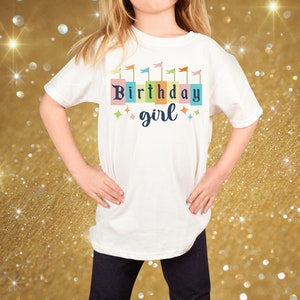 Peut inclure: T-shirt blanc avec un motif d'anniversaire fille color&eacute;. Le motif pr&eacute;sente une rang&eacute;e de drapeaux color&eacute;s et les mots "Birthday girl" dans une police de caract&egrave;res en blocs color&eacute;s.