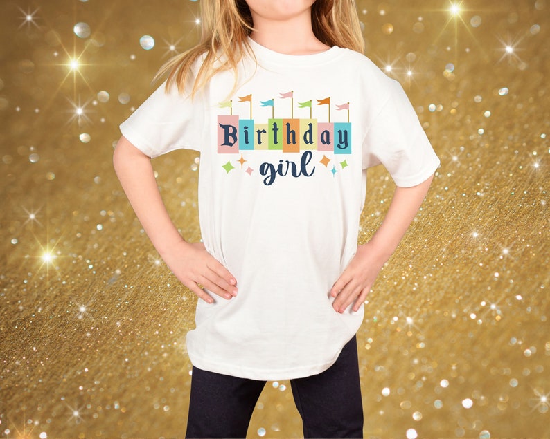 Peut inclure: T-shirt blanc avec un motif d'anniversaire fille color&eacute;. Le motif pr&eacute;sente une rang&eacute;e de drapeaux color&eacute;s et les mots "Birthday girl" dans une police de caract&egrave;res en blocs color&eacute;s.
