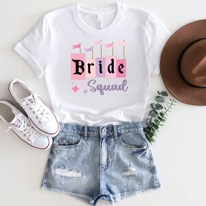 May include: White t-shirt with a pink and purple graphic that says "Bride Squad" with a castle design. The shirt is paired with denim shorts and white sneakers.