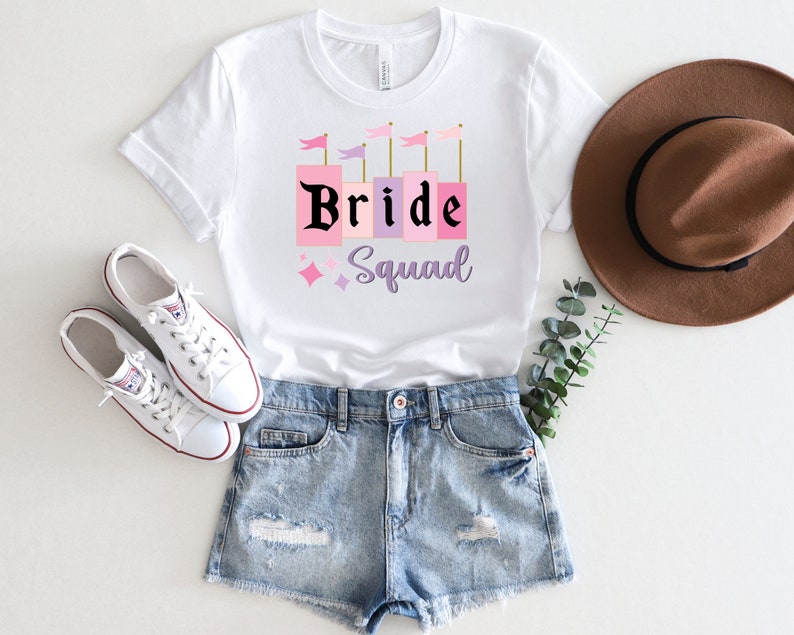 May include: White t-shirt with a pink and purple graphic that says "Bride Squad" with a castle design. The shirt is paired with denim shorts and white sneakers.
