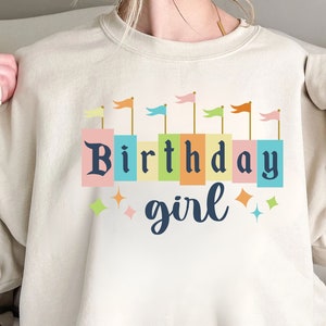 Peut inclure: Un sweat-shirt beige avec un motif graphique de drapeaux color&eacute;s et le texte "Birthday girl".