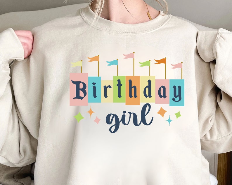 Peut inclure: Un sweat-shirt beige avec un motif graphique de drapeaux color&eacute;s et le texte "Birthday girl".