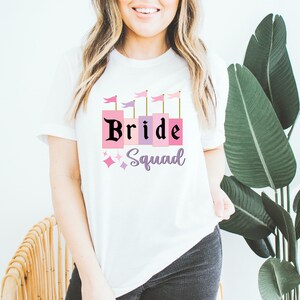 May include: White t-shirt with a pink and purple castle graphic and the text "Bride Squad" in black.