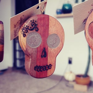 May include: Two wooden skull ornaments with coins embedded in the eye sockets. The skulls are hanging from brown cords and have a dark wood stain. The text on the tag reads "Suertes Sin Limites".