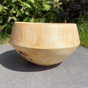 May include: A light-coloured wooden bowl with a natural finish. The bowl has a wide, shallow design with a slightly angled rim. The wood grain is visible, adding to the bowl's rustic charm. Ideal for serving or as a decorative piece.