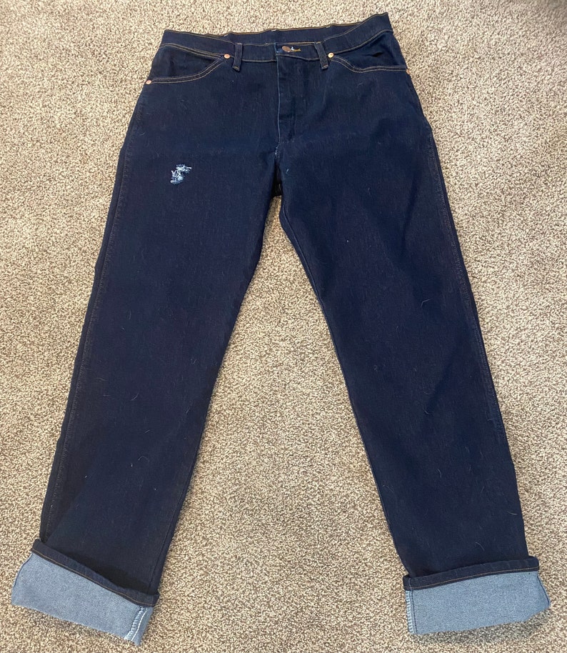 May include: A pair of dark blue denim jeans with cuffed hems. The jeans have a distressed look with a small hole on the left leg.