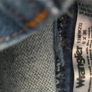 May include: A white tag with black text that reads "Wrangler 13MWZG 5 X 36". The tag is attached to a pair of blue jeans. The text on the tag instructs to machine wash warm with like colours.