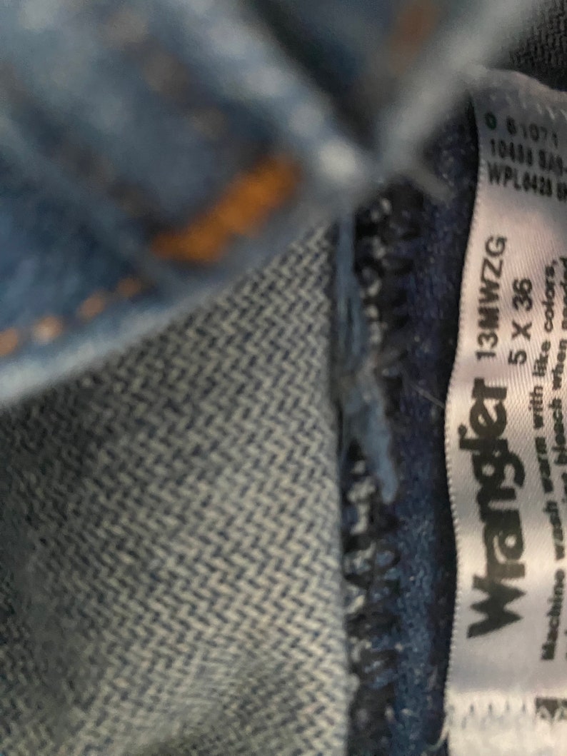 May include: A white tag with black text that reads "Wrangler 13MWZG 5 X 36". The tag is attached to a pair of blue jeans. The text on the tag instructs to machine wash warm with like colours.