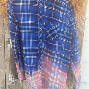 May include: Blue plaid button-down shirt with a bleached, ombre effect. The shirt features a classic collar, a single chest pocket, and long sleeves. The bottom half of the shirt fades from blue to pink and orange.