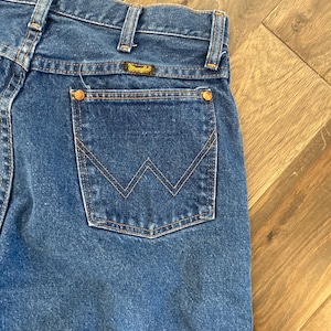 K&ouml;nnte beinhalten: Nahaufnahme einer blauen Jeans mit klassischem F&uuml;nf-Taschen-Design. Die Ges&auml;&szlig;tasche hat ein markantes, aufgesticktes 'W'-Muster. Die Jeans hat ein Wrangler-Markenetikett und kupferfarbene Nieten. Der Denim hat eine mittlere Waschung.