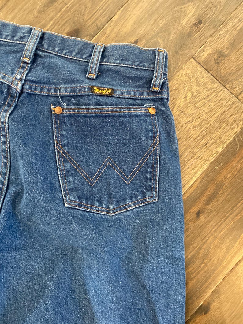 K&ouml;nnte beinhalten: Nahaufnahme einer blauen Jeans mit klassischem F&uuml;nf-Taschen-Design. Die Ges&auml;&szlig;tasche hat ein markantes, aufgesticktes 'W'-Muster. Die Jeans hat ein Wrangler-Markenetikett und kupferfarbene Nieten. Der Denim hat eine mittlere Waschung.