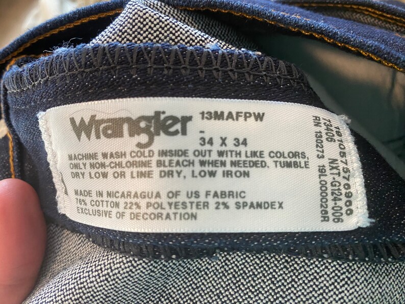 May include: A white Wrangler jeans care label with black text. The label says "13MAFPW - 34 X 34" and includes washing instructions. The label also says "Made in Nicaragua of US Fabric 76% Cotton 22% Polyester 2% Spandex Exclusive of Decoration."