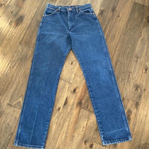 K&ouml;nnte beinhalten: Eine blaue Jeans mit klassischem geradem Bein. Die Jeans hat einen Knopf- und Rei&szlig;verschluss, Vorder- und Ges&auml;&szlig;taschen und besteht aus einem mittelblauen Denim-Material. Die Jeans liegt flach auf einer Holzoberfl&auml;che.