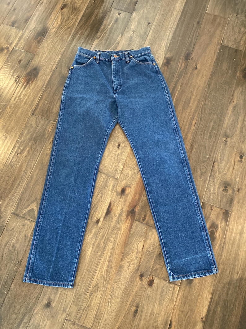 K&ouml;nnte beinhalten: Eine blaue Jeans mit klassischem geradem Bein. Die Jeans hat einen Knopf- und Rei&szlig;verschluss, Vorder- und Ges&auml;&szlig;taschen und besteht aus einem mittelblauen Denim-Material. Die Jeans liegt flach auf einer Holzoberfl&auml;che.