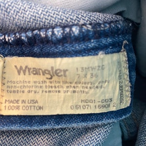 May include: Close-up of a Wrangler jeans label. The label is off-white with black text, including the brand name, size 7x36, and care instructions. The jeans are made of 100% cotton and made in the USA.