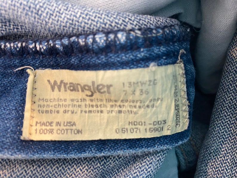 May include: Close-up of a Wrangler jeans label. The label is off-white with black text, including the brand name, size 7x36, and care instructions. The jeans are made of 100% cotton and made in the USA.
